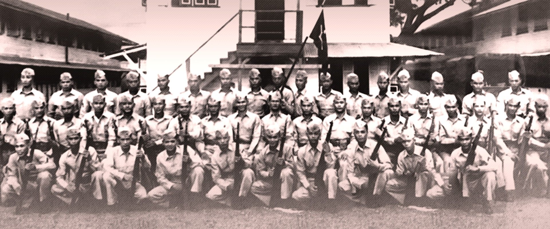 A black and white photograph of a group of uniformed individuals posing in front of a building with multiple steps leading up to it. The individuals are arranged in three rows, with those in the front row seated and holding rifles. The uniforms consist of shirts, ties, and caps. The background features the building and a tree.