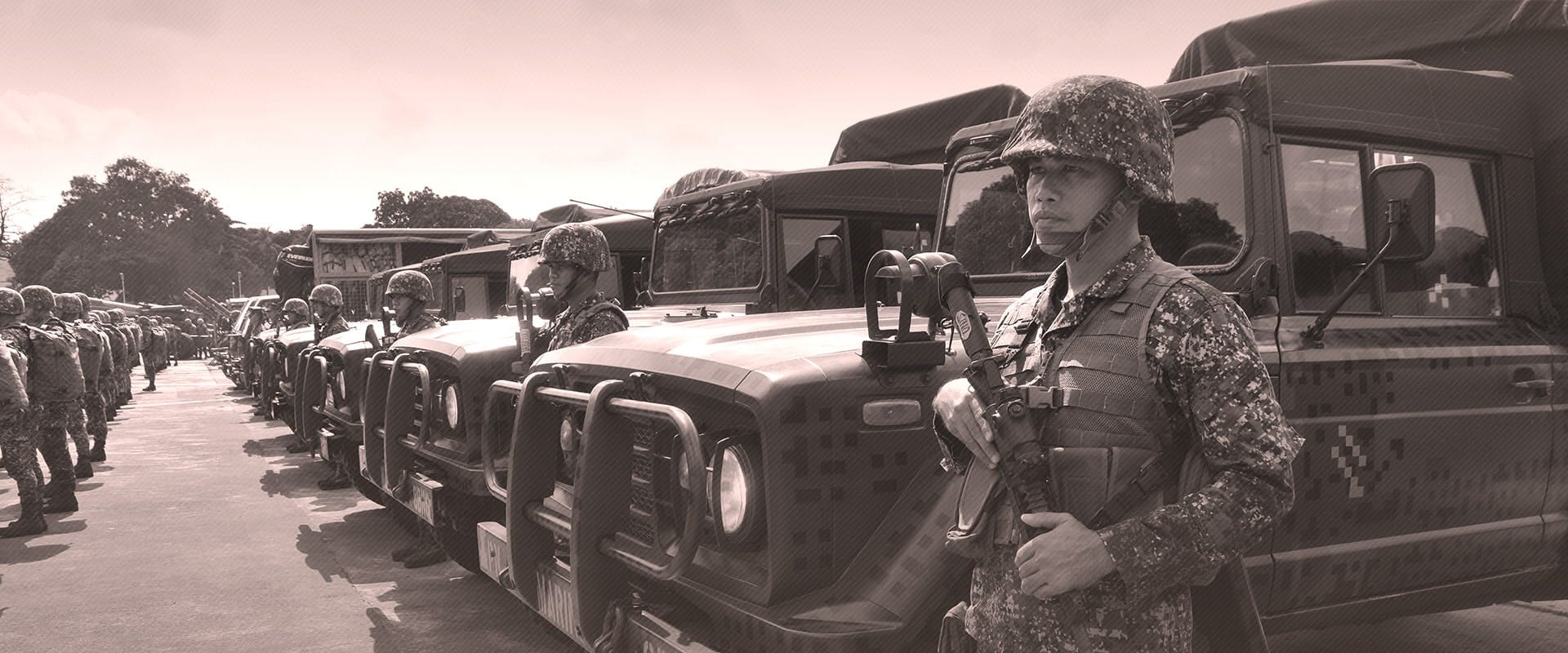A grayscale photograph of a line of military personnel standing next to armored vehicles. The soldier in the foreground, wearing a camouflaged uniform and helmet, holds a communication device. Behind him, rows of soldiers stand at attention, and multiple military vehicles are parked, all under a partly cloudy sky.