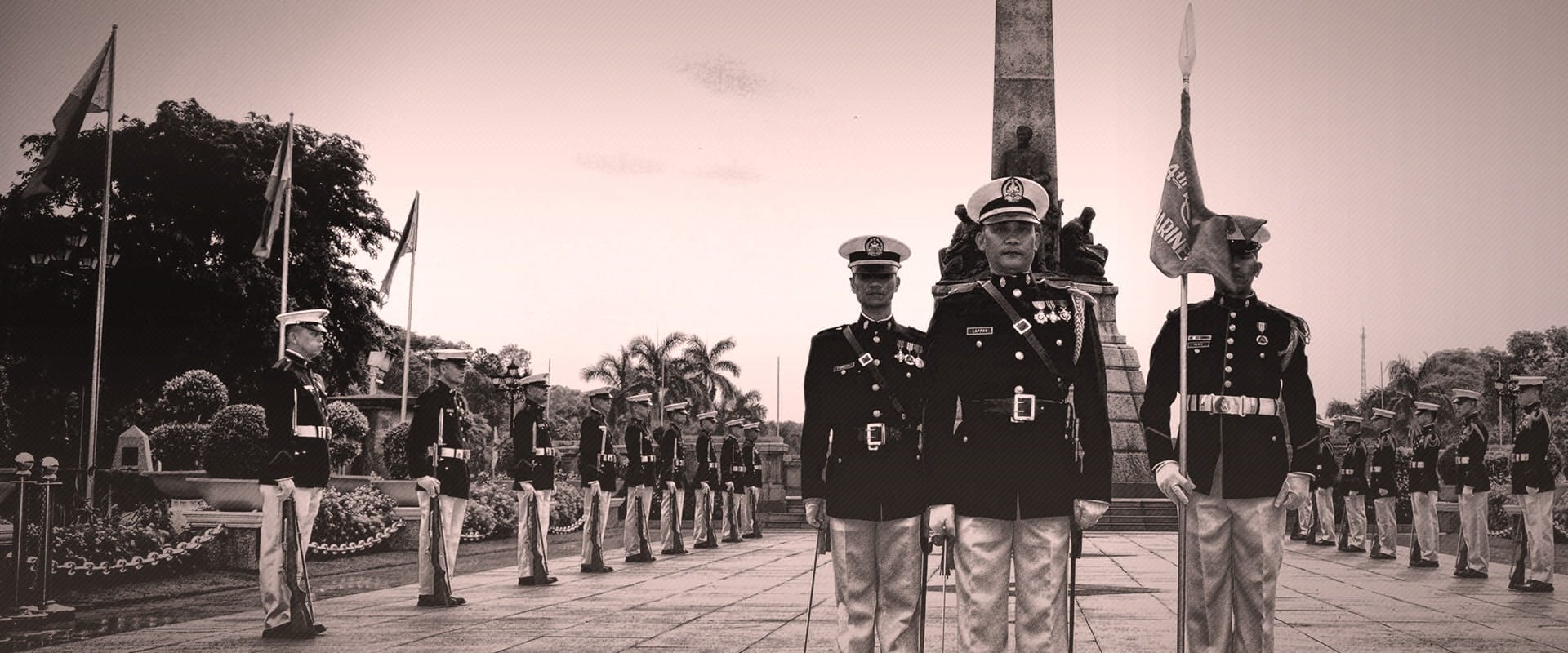 Black and white photo of a group of military personnel in full ceremonial uniforms, standing in formation near a monument with flags. They are surrounded by lush greenery, and the sky is lightly clouded.